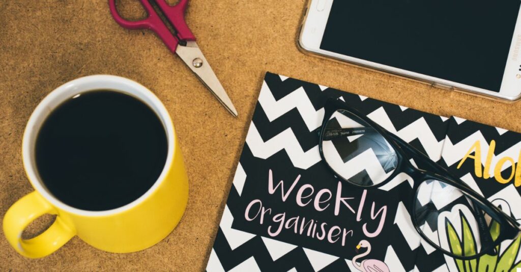 Imagen de una taza de café, un cuaderno con 'Weekly Organizer', un teléfono y unas tijeras en un entorno de escritorio, ilustrando herramientas para una mejor organización del espacio virtual.