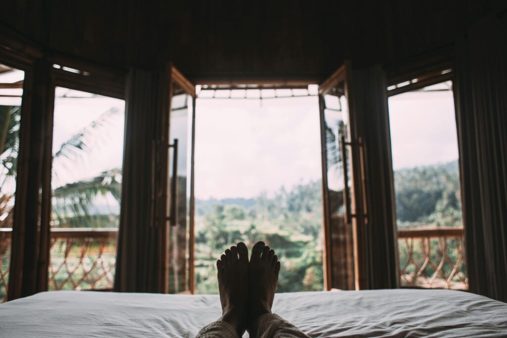 Pies descansando sobre una cama con vista a un paisaje verde, evocando comodidad y serenidad.